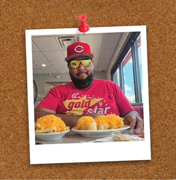 Person seated at a table with 3 cheese coneys and a plate of cheese fries